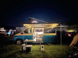 A blue and white two toned campervan on a campsite adorned with fairy lights and an outdoor table set up
