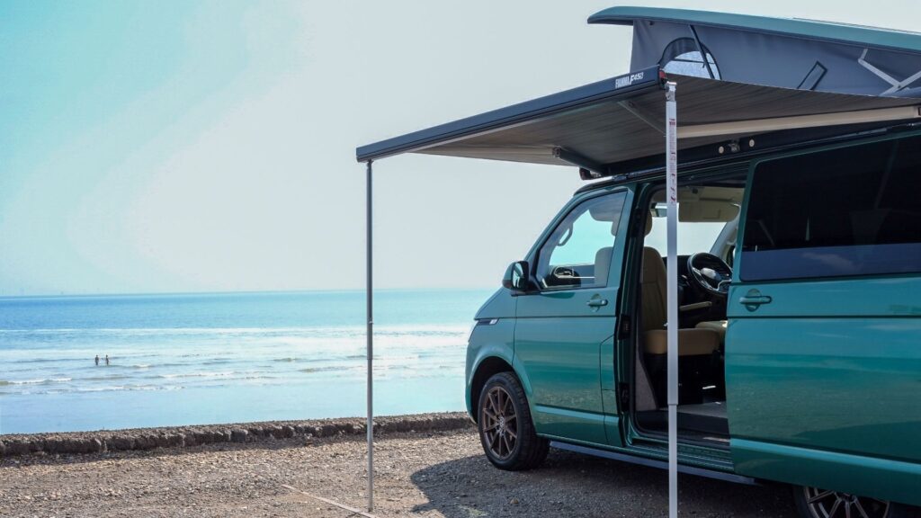 a green campervan with awning and pop-top along a beach road overlooking the sea with swimmers